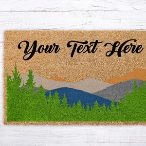 May include: A rectangular doormat with a mountain landscape design. The mat features a tan background with a green forest at the bottom, and blue, grey, and orange mountains. The text "Your Text Here" is written across the top in black script.
