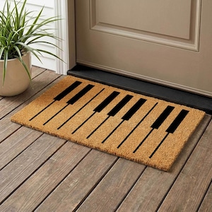 May include: A rectangular doormat featuring a piano key design. The mat is a natural tan colour with black piano keys. The mat is placed on a wooden porch.