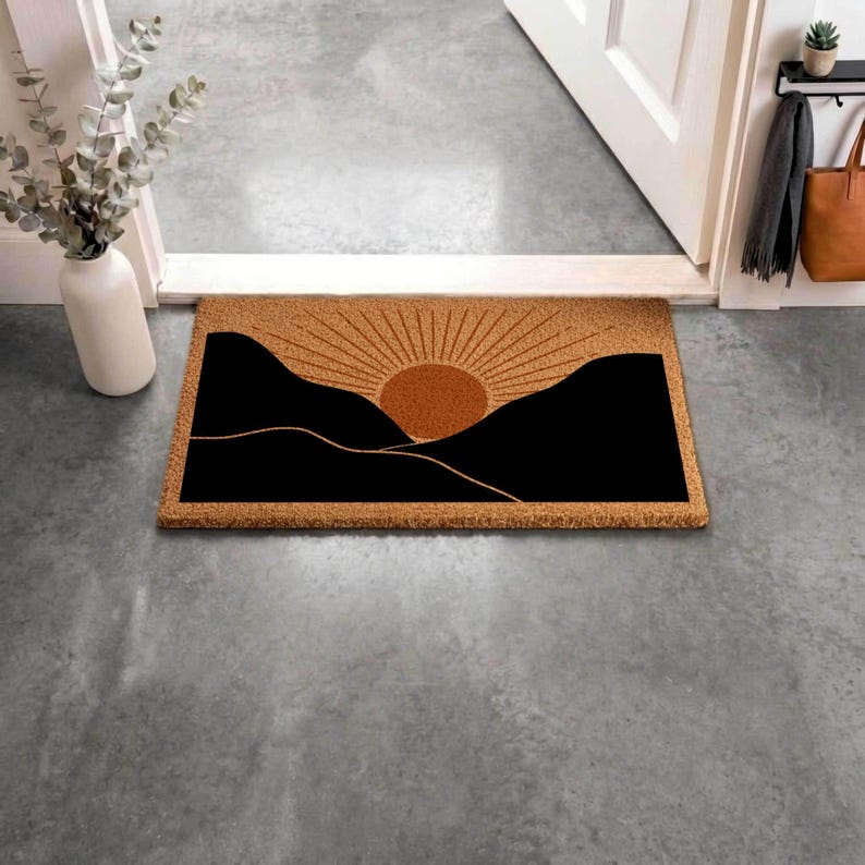 May include: Rectangular doormat with a sunrise design. The mat features a brown background with a black mountain silhouette and a sun with radiating lines. Placed in front of a white door, the mat adds a decorative touch to the entryway.