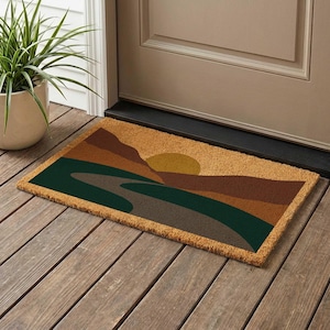 May include: Rectangular doormat with a landscape design. The mat features a sunset over mountains in shades of brown, green, and yellow. The mat is made of a natural fiber material and is placed on a wooden porch.