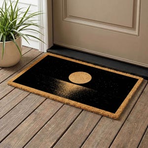 A black and gold door mat with a design depicting a sunset or sunrise on a wooden floor next to a door.