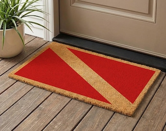 May include: Rectangular doormat featuring a red and tan scuba diving flag design. The mat is made of natural fibres and is placed on a wooden porch. The red sections are triangular, with a tan diagonal stripe.