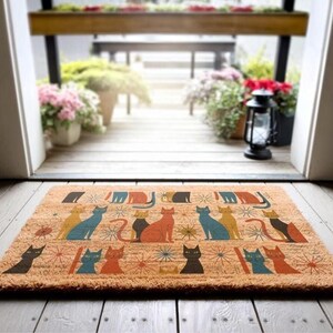 A colorful, patterned door mat with various cat illustrations is placed on a wooden floor, with a view of a wooden deck and potted plants through an open doorway.