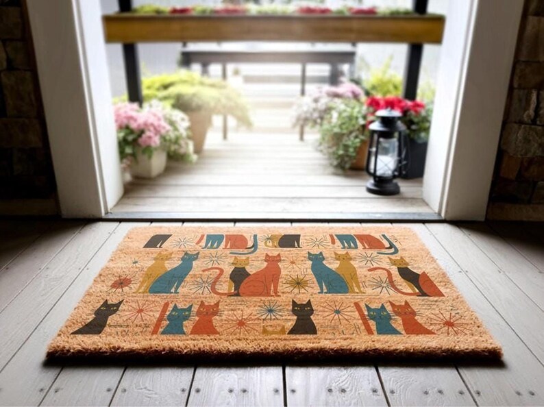 A colorful, patterned door mat with various cat illustrations is placed on a wooden floor, with a view of a wooden deck and potted plants through an open doorway.