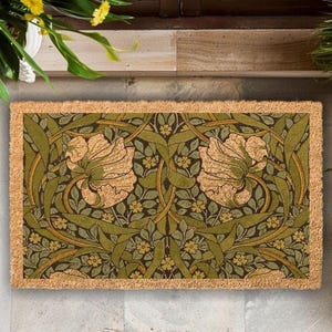 A decorative door mat with a floral pattern in shades of green and brown, placed on a wooden floor next to a window with plants.