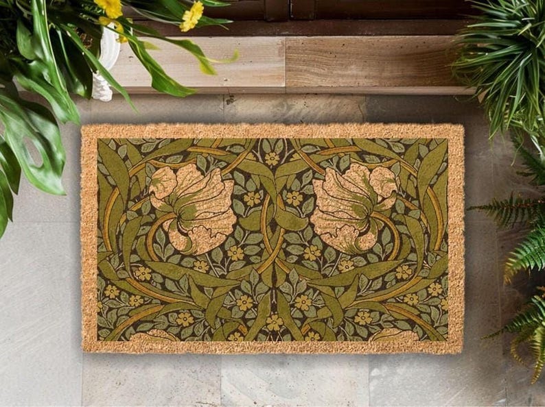 A decorative door mat with a floral pattern in shades of green and brown, placed on a wooden floor next to a window with plants.