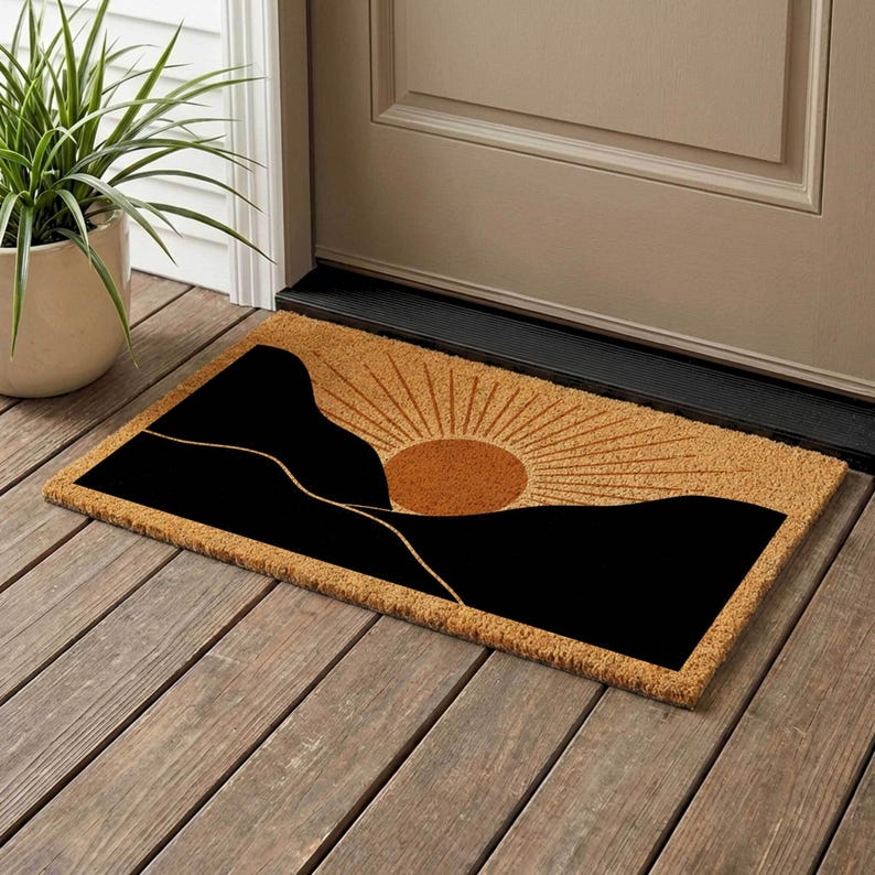 May include: A rectangular doormat with a black mountain silhouette design and a rising sun graphic. The sun is a warm brown color with radiating lines. The mat is made of a natural fiber material.