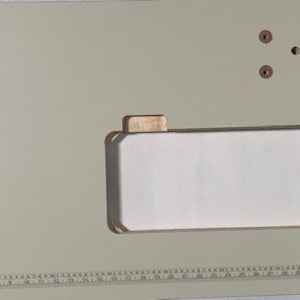 May include: A light green sewing machine table with a white rectangular sewing machine bed. The table has a ruler along the bottom edge and several small holes in the surface.