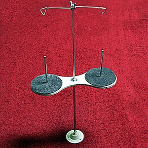 May include: A silver metal thread stand with two spools and a rotating arm. The stand has a black base with a white center.