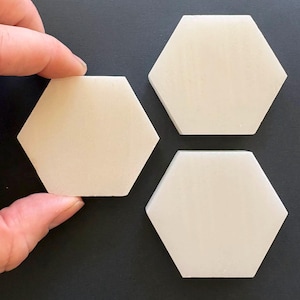 Puede incluir: Tres baldosas hexagonales blanquecinas sobre una superficie oscura. Una mano sostiene una baldosa. Las baldosas tienen un acabado liso y mate. La imagen está tomada desde arriba.