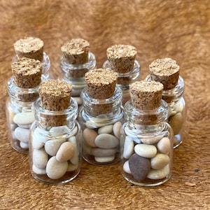 Puede incluir: Ocho pequeñas botellas de vidrio con tapas de corcho llenas de piedras lisas de color marrón claro, gris y blanco.