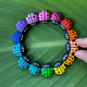 May include: A circle of painted ladybirds in various colours, including red, orange, yellow, green, blue, purple, and pink. Each ladybird has black spots and a black face with white eyes.