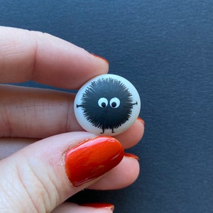 Puede incluir: Un botón pequeño, redondo y blanco con una criatura negra y peluda con ojos grandes y pequeñas patas pintadas en él.