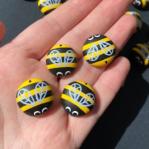 May include: A hand holding six painted bee-themed rocks. The rocks are painted black and yellow with white wings and black eyes.