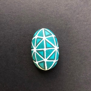 Pode incluir: Uma pedra pintada em azul-turquesa e branco com um padrão geométrico de triângulos e linhas.