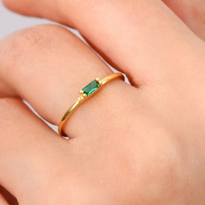 May include: A gold ring with a rectangular emerald-green gemstone. The ring is thin and delicate, with a simple band. The gemstone is set horizontally in the center of the ring.