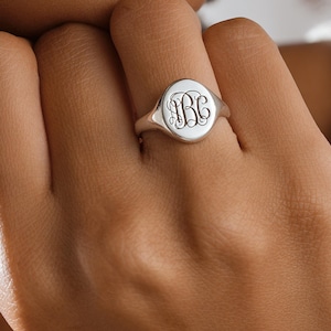 May include: A silver signet ring with a monogram design. The ring has a round face with an engraved monogram. The ring is worn on a finger. The ring is a classic piece of jewelry.