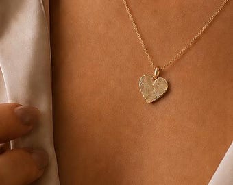 Könnte beinhalten: Eine goldene herzförmige Anhängerkette an einer feinen Kette. Der Anhänger zeigt auf einer Seite eine Fingerabdruckstruktur und auf der Rückseite den eingravierten Text "Forever in my heart" mit einem kleinen Herzsymbol, dargestellt in zwei Insets.