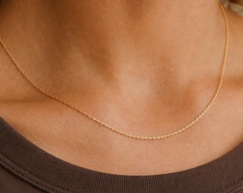 May include: A delicate gold chain necklace featuring a fine, interlocking link design. The necklace rests against the neck, showcasing a polished metallic finish that catches the light. The simple, slender construction provides a subtle, refined appearance.