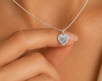 Puede incluir: Un collar de plata con un pequeño colgante en forma de corazón que presenta una textura de huella dactilar detallada en su superficie. El colgante cuelga de una delicada cadena de plata. La palabra LÖVINKA aparece en la esquina inferior derecha.
