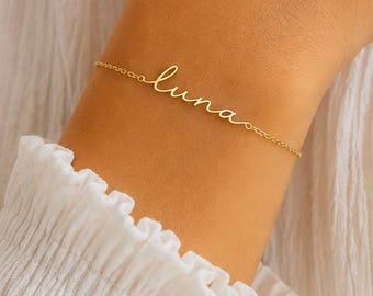 May include: A gold-toned metal chain bracelet featuring the name "luna" written in a delicate, cursive script font. The bracelet is secured with a thin, matching gold-toned metal chain that wraps around the wrist, showcasing a minimalist design.