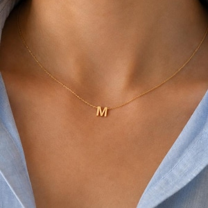 May include: A delicate gold-toned necklace featuring a single, centered capital letter M pendant. The pendant is suspended from a thin, fine-link chain that rests against the collarbone, creating a simple and refined look for everyday wear.