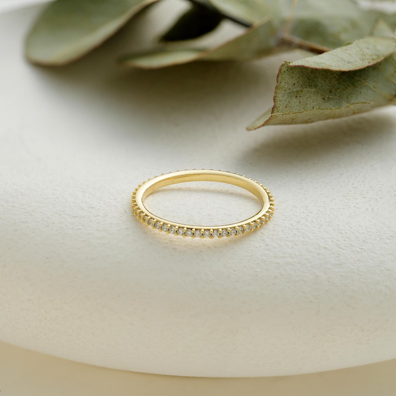 May include: A delicate gold ring with a continuous row of small, sparkling gemstones. The ring is set against a soft, white background, with a sprig of green leaves in the background. The ring's design is simple and elegant.
