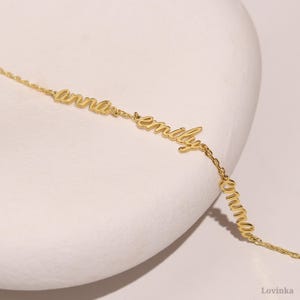May include: A delicate gold-colored necklace featuring the names "anna", "emily", and "emma" in a cursive font. The names are connected by a fine chain. The jewelry is displayed on a white surface.