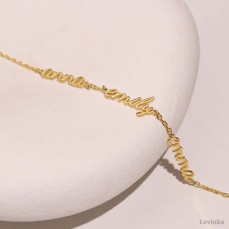 May include: A delicate gold-colored necklace featuring the names "anna", "emily", and "emma" in a cursive font. The names are connected by a fine chain. The jewelry is displayed on a white surface.