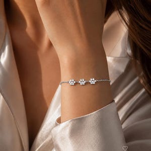 May include: A silver-toned chain bracelet featuring three small paw print charms, each engraved with a name. The bracelet is delicate and minimalist in design, with a secure clasp. The text LOVINKA and a small heart icon appear in the bottom right corner.
