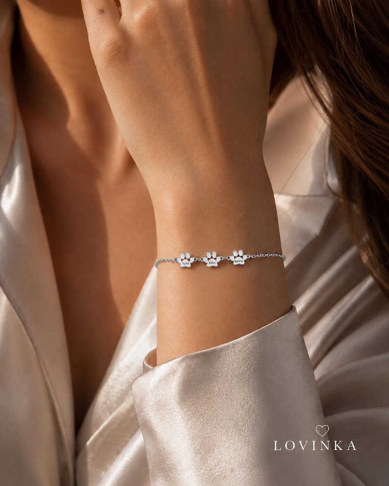 May include: A silver-toned chain bracelet featuring three small paw print charms, each engraved with a name. The bracelet is delicate and minimalist in design, with a secure clasp. The text LOVINKA and a small heart icon appear in the bottom right corner.
