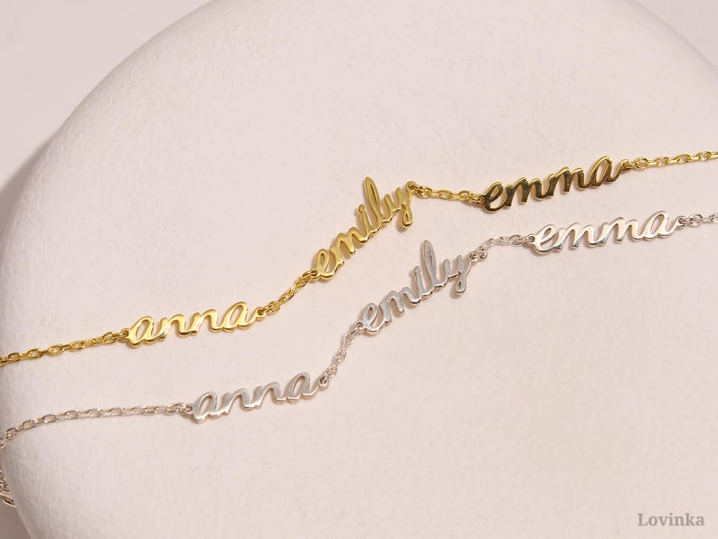 May include: Three delicate name necklaces in gold and silver. The names "Anna", "Emily", and "Emma" are displayed in a cursive font. The necklaces are on a white surface.