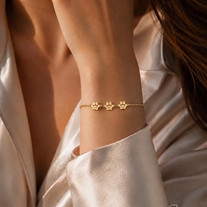 May include: A gold-toned chain bracelet featuring three small, evenly spaced paw print charms. Each charm is engraved with a name. The bracelet is delicate, with a fine link chain design that secures around the wrist, showcasing a polished finish.