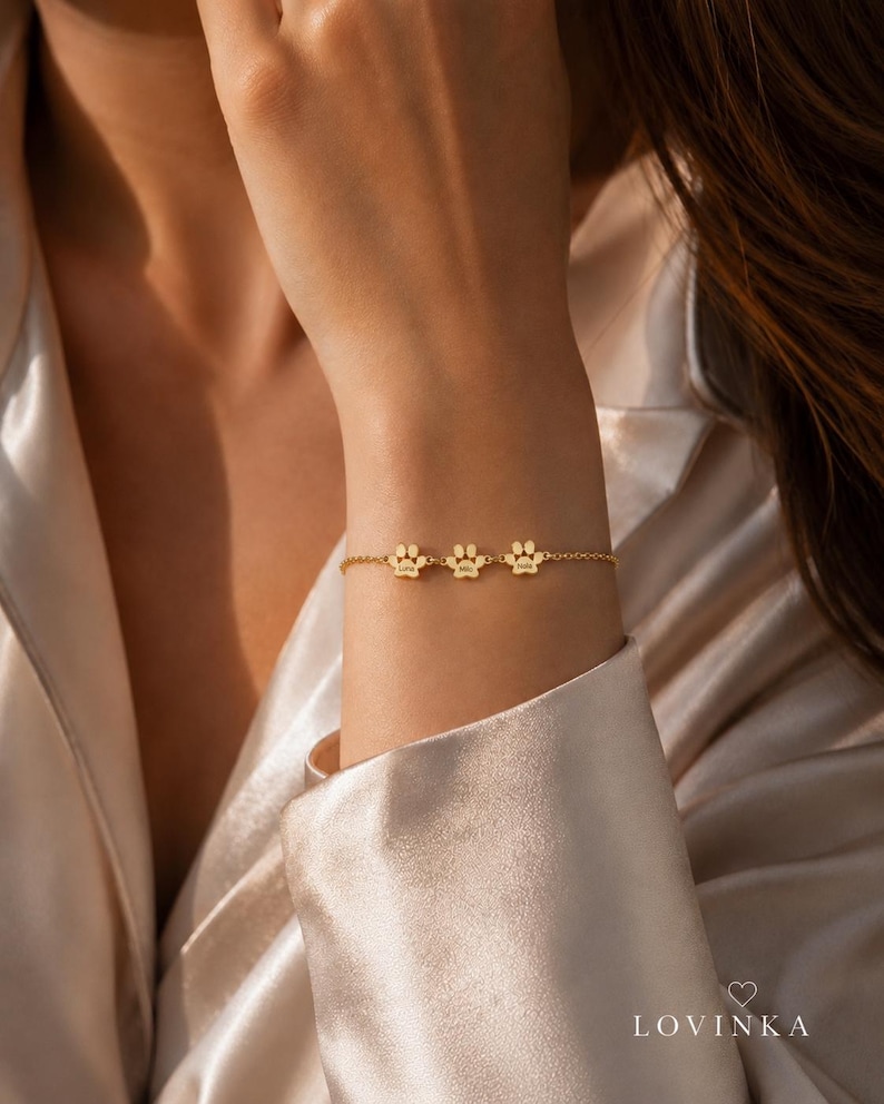 May include: A gold-toned chain bracelet featuring three small, evenly spaced paw print charms. Each charm is engraved with a name. The bracelet is delicate, with a fine link chain design that secures around the wrist, showcasing a polished finish.