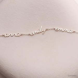 May include: A delicate silver necklace featuring the names "anna", "emily", and "emma" in a cursive font. The names are connected by a fine chain, creating a personalized jewelry piece. The necklace is displayed on a neutral background.