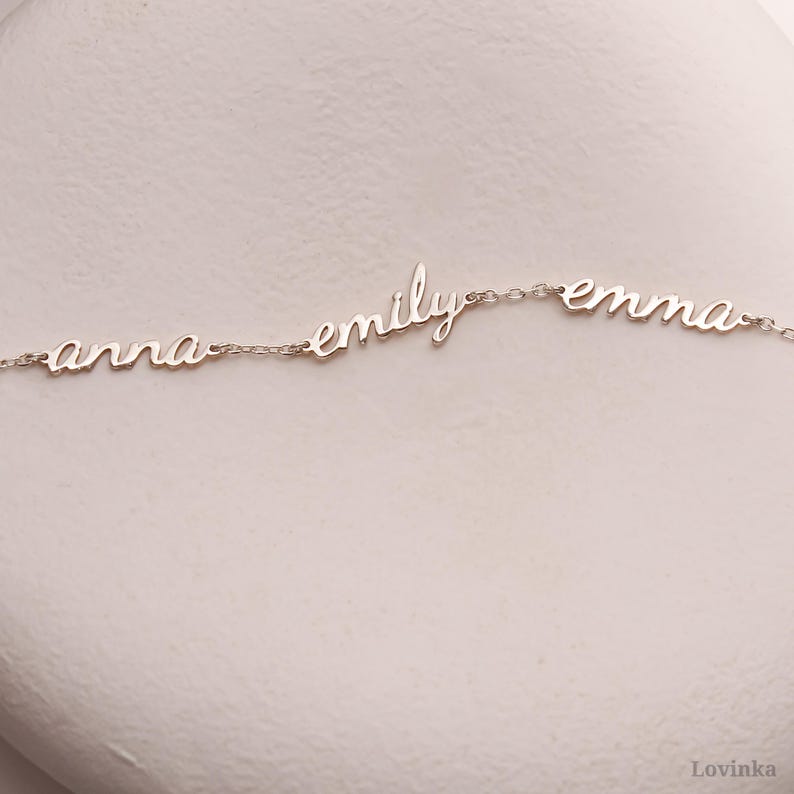 May include: A delicate silver necklace featuring the names "anna", "emily", and "emma" in a cursive font. The names are connected by a fine chain, creating a personalized jewelry piece. The necklace is displayed on a neutral background.
