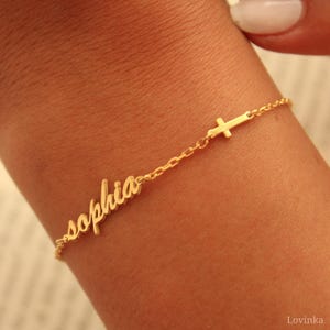 May include: A delicate gold bracelet featuring the name "Sophia" in cursive script and a small cross charm. The bracelet is attached to a fine chain, creating an elegant and personalized accessory.