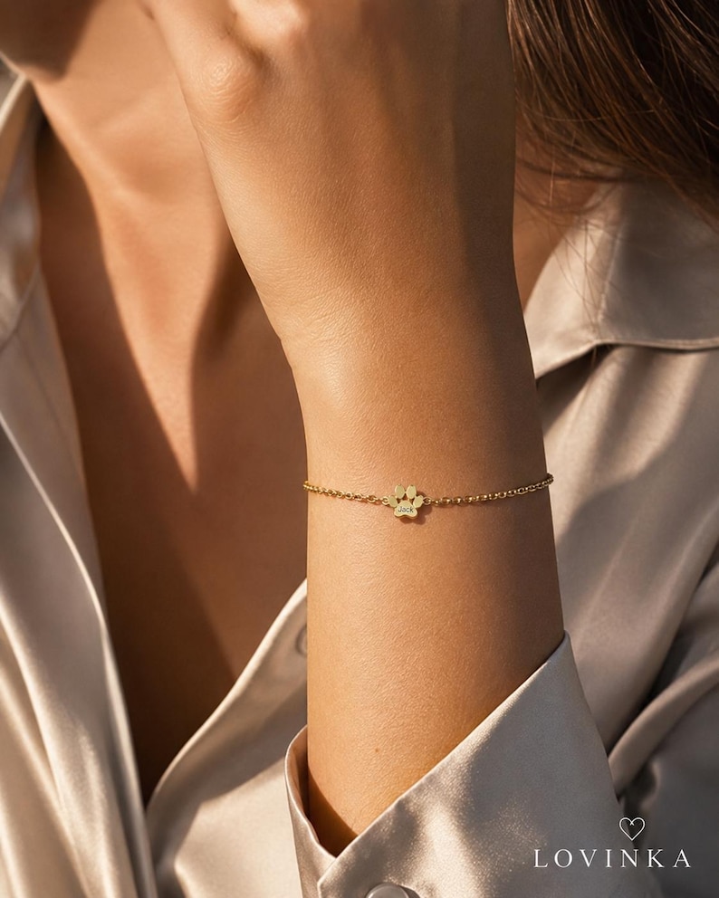 May include: A gold-toned chain bracelet featuring a small, central paw print charm. The charm is engraved with the name Jack. The bracelet is constructed with a delicate, fine link chain that secures around the wrist with a simple clasp mechanism.