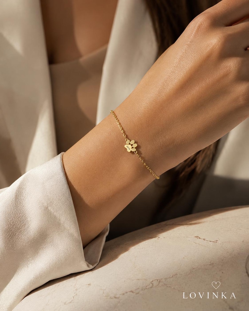 May include: A delicate gold-toned chain bracelet featuring a small, central paw print charm. The charm is engraved with the name Jack. The bracelet consists of thin, interlocking circular links and secures with a simple clasp mechanism.