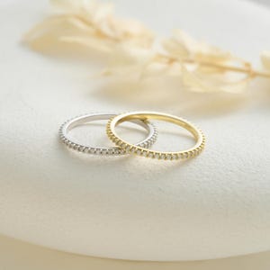 May include: Two delicate rings, one silver and one gold, are displayed. Both rings are adorned with small, sparkling stones set around the band. The rings are positioned on a white surface, with soft, blurred foliage in the background, creating a simple, elegant composition.
