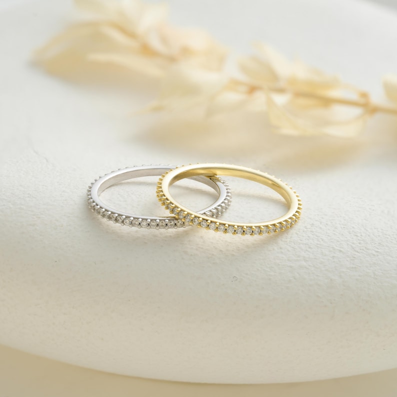 May include: Two delicate rings, one silver and one gold, are displayed. Both rings are adorned with small, sparkling stones set around the band. The rings are positioned on a white surface, with soft, blurred foliage in the background, creating a simple, elegant composition.