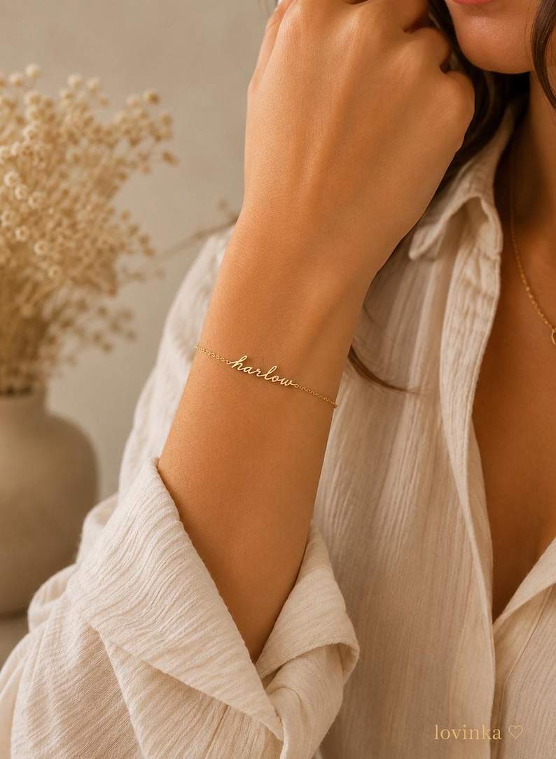 K&ouml;nnte beinhalten: Ein zartes, goldfarbenes Kettenarmband mit einer mittigen Namensplatte, auf der das Wort "harlow" in einer kursiven Schreibschrift steht. Das Armband wird durch eine d&uuml;nne, polierte Metallkette am Handgelenk gehalten.