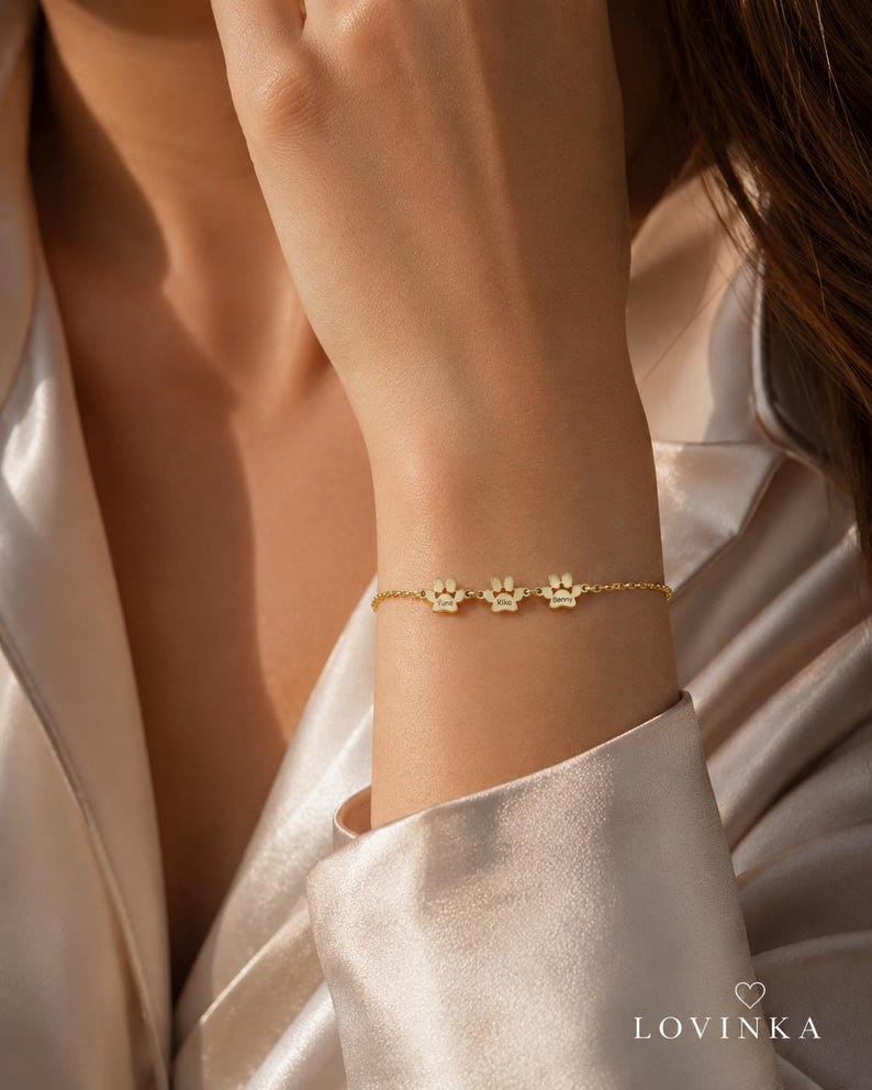 May include: A gold-toned chain bracelet featuring three small, evenly spaced paw print charms. Each charm is engraved with a name. The bracelet is secured with a delicate link chain. The text LOVINKA with a heart icon is visible in the corner.