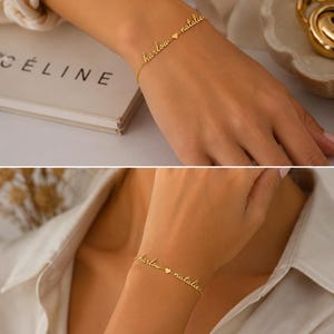 K&ouml;nnte beinhalten: Ein goldfarbenes Kettenarmband mit zwei in Schreibschrift verfassten Namen, "harlow" und "natalie", die durch einen kleinen herzf&ouml;rmigen Anh&auml;nger getrennt sind. Die feine Metallkette verbindet die Namen zu einem durchgehenden, personalisierten Schmuckst&uuml;ck.