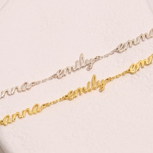 May include: Two delicate name bracelets. One is silver-toned, spelling "emily" and "emma". The other is gold-toned, spelling "anna" and "emily". The bracelets are on a white surface.