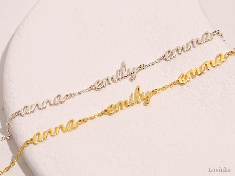 May include: Two delicate name bracelets. One is silver-toned, spelling "emily" and "emma". The other is gold-toned, spelling "anna" and "emily". The bracelets are on a white surface.