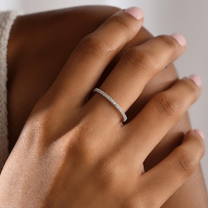 May include: A delicate, silver-toned ring with a continuous row of small, clear gemstones. The ring is shown on a person's finger, highlighting the sparkle of the stones. The ring is a classic design, suitable for everyday wear or special occasions.