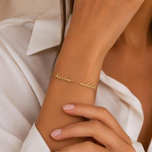 K&ouml;nnte beinhalten: Ein goldfarbenes Metallkettenarmband mit den zwei Namen "harlow" und "natalie" in einer kursiven Schreibschrift. Die Namen sind direkt in die feine Gliederkette eingearbeitet und bilden ein durchgehendes, personalisiertes Schmuckst&uuml;ck.