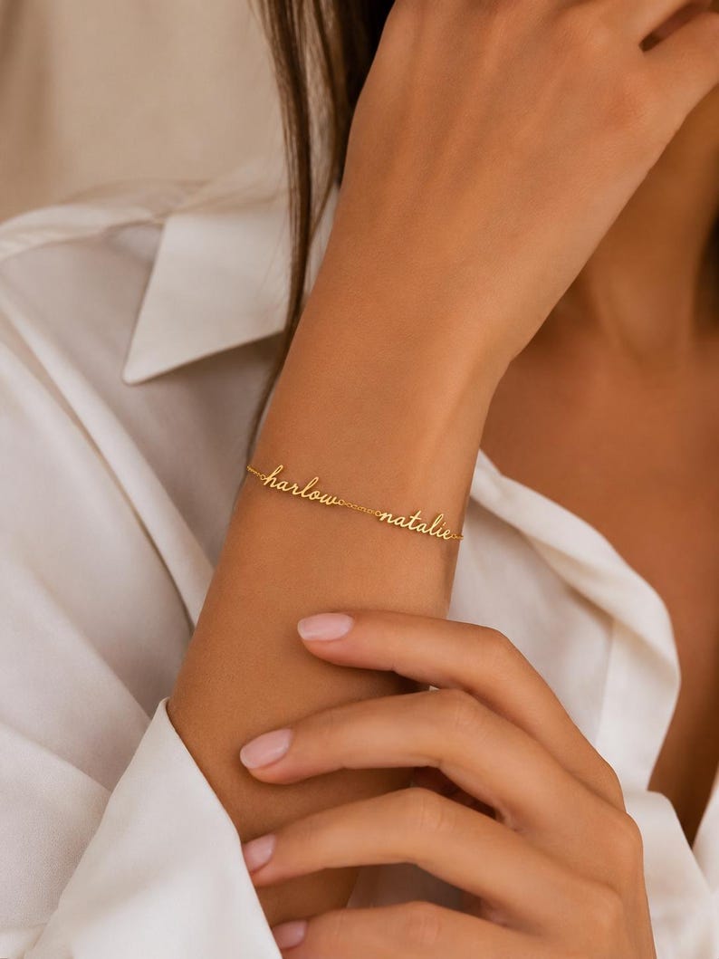 K&ouml;nnte beinhalten: Ein goldfarbenes Metallkettenarmband mit den zwei Namen "harlow" und "natalie" in einer kursiven Schreibschrift. Die Namen sind direkt in die feine Gliederkette eingearbeitet und bilden ein durchgehendes, personalisiertes Schmuckst&uuml;ck.