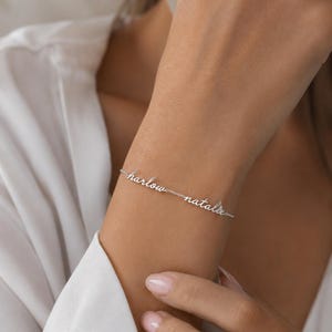 K&ouml;nnte beinhalten: Ein silberfarbenes Kettenarmband mit den zwei Namen Harlow und Natalie in kursiver Schreibschrift. Die Namen sind durch eine feine Metallkette miteinander verbunden und bilden ein durchgehendes Schmuckst&uuml;ck auf der Haut.